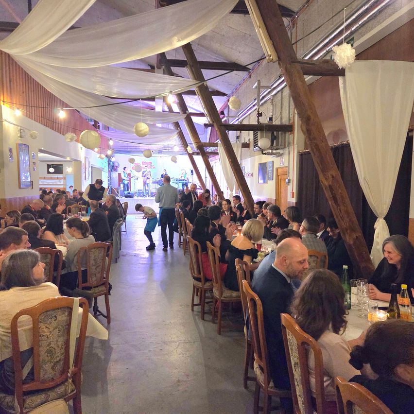A large hall with tables and chairs where people are sitting and eating. A person is walking on the floor, and a few people are standing. There is a stage in the middle with a band playing.