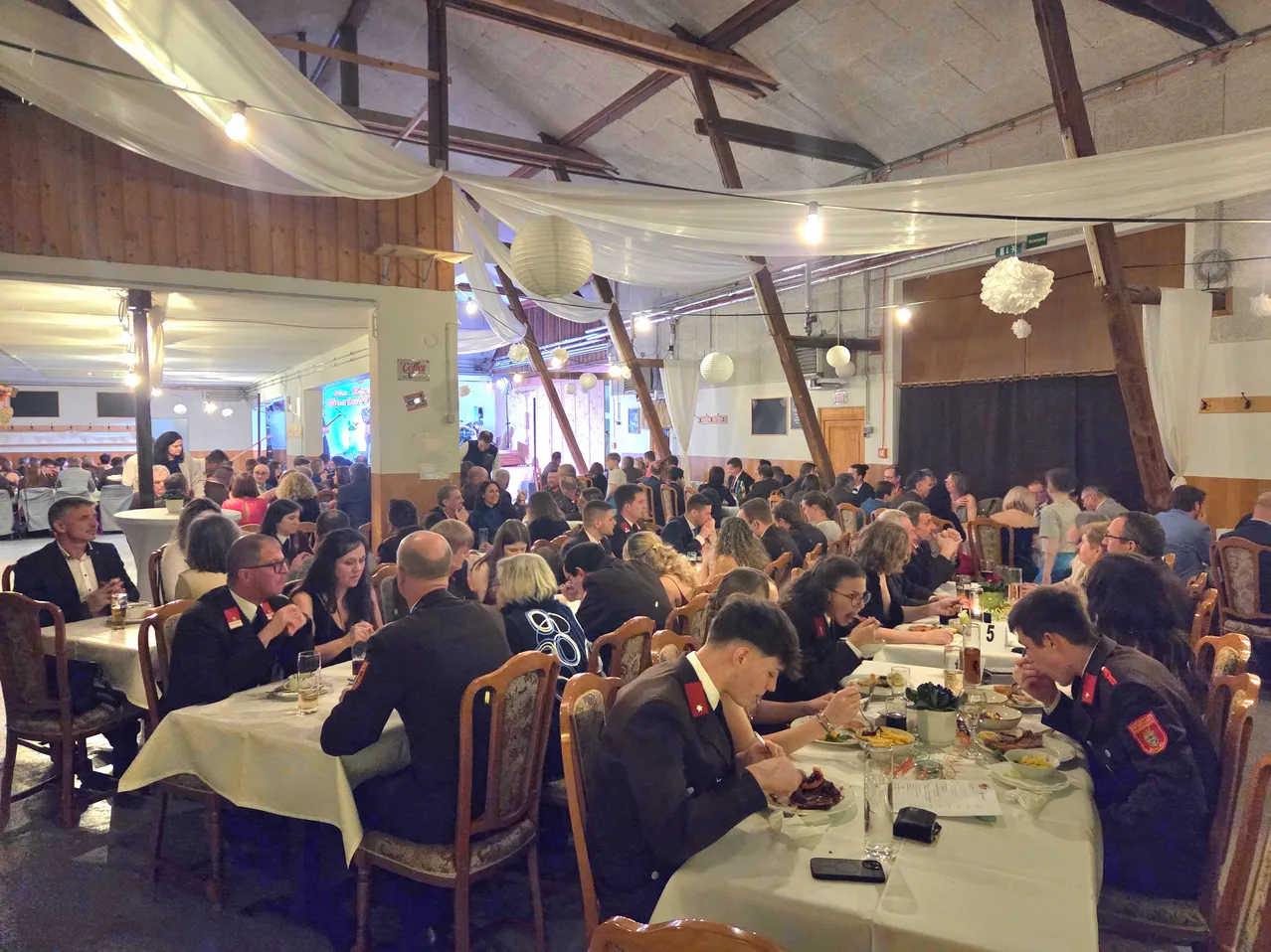 Ein großer Saal mit Holzbalken und weißen Vorhängen verfügt über mehrere lange Esstische. Zahlreiche Menschen in formeller Kleidung, einschließlich Militäruniformen, sind anwesend und nehmen Mahlzeiten ein. Die Umgebung ist warm mit hängenden Lampen und dekorativen Elementen.