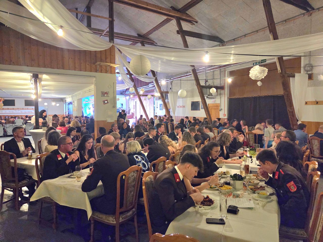 A large hall with wooden beams and white curtains features multiple long dining tables. Numerous people in formal attire, including military uniforms, are seated, engaging in meals. The setting is warm with hanging lamps and decorative elements.