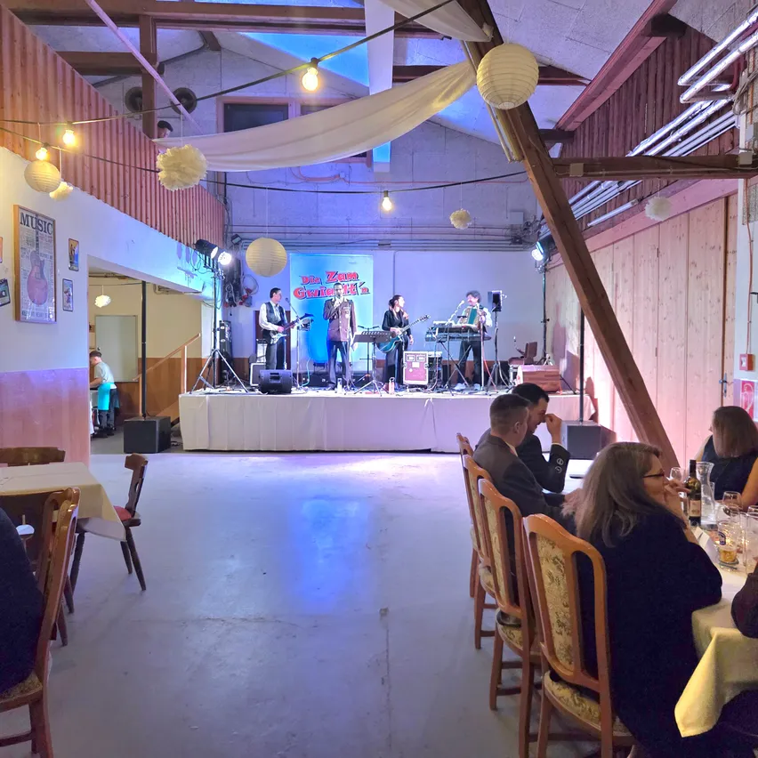 Eine Band spielt auf einer Bühne in einem Restaurant, während mehrere Personen an Tischen sitzen und zuschauen. Die Bühne ist mit einem weißen Tuch bedeckt und hat Musikinstrumente und Lichter.