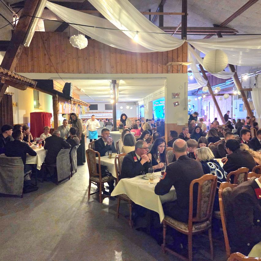 A large group of people are seated at tables in a dining room with wooden walls and ceiling. There are white drapes and paper lanterns hanging above.