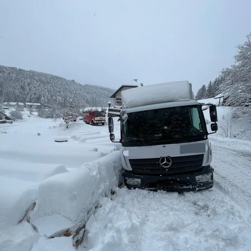 Ein Mercedes-Lkw ist in tiefem Schnee auf einer verschneiten Straße steckengeblieben, mit einem roten Feuerwehrwagen in der Nähe. Bäume und Häuser sind im Hintergrund sichtbar.