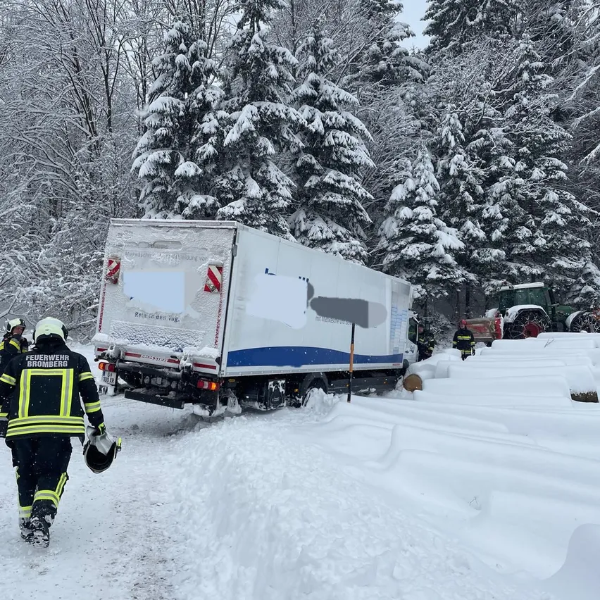 Feuerwehrleute navigieren eine verschneite Straße mit einem festsitzenden LKW am Straßenrand, umgeben von schneebedeckten Bäumen und einem Traktor in der Nähe.