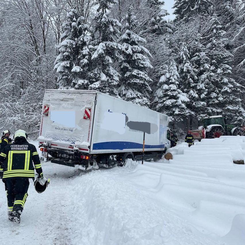 Firefighters are navigating a snowy road with a truck stuck on the side, amidst snow-covered trees and a tractor nearby.