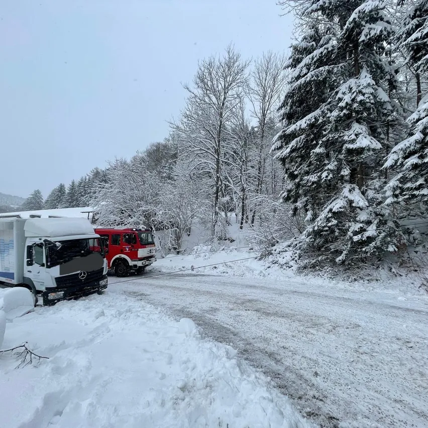 Zwei Lastwagen sind auf einer verschneiten Straße geparkt, umgeben von schneebedeckten Bäumen. Das Gebiet scheint ein Gebirgsregion zu sein.