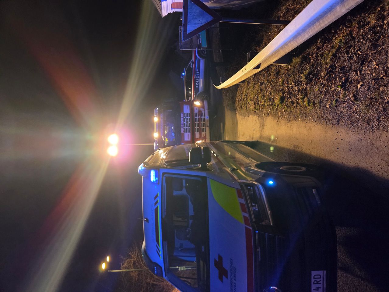 Ein Krankenwagen mit eingeschaltetem Licht steht nachts auf einer Straße. Dahinter befinden sich ein Polizeiwagen und ein weiteres Notfallfahrzeug.