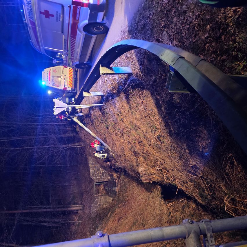 Ein Krankenwagen mit blinkenden Lichtern steht am Straßenrand, und Rettungskräfte kümmern sich um eine Person am Boden in der Nähe einer Leitplanke.