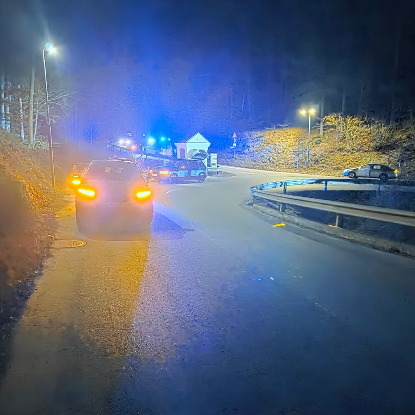 In der Nacht fahren mehrere Autos auf einer kurvenreichen Straße mit Straßenlaternen und einem Geländer. Ein Pickup-Truck und ein Auto sind vor ihnen angehalten.