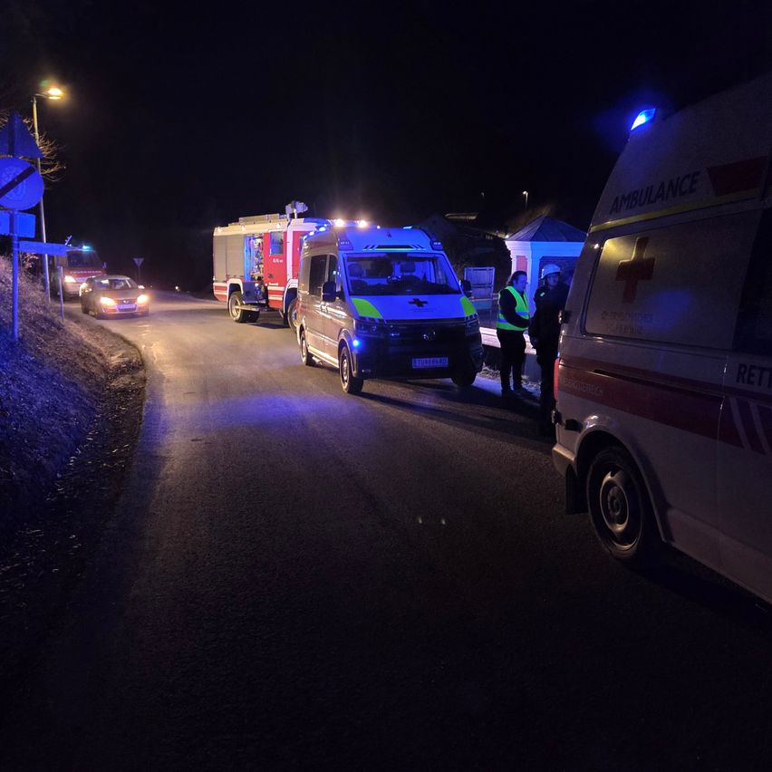 Eine Notfallszene bei Nacht, mit mehreren Fahrzeugen auf der Straße. Ein Krankenwagen, ein Feuerwehrwagen und ein Polizeiauto sind geparkt. Rettungskräfte sind anwesend.
