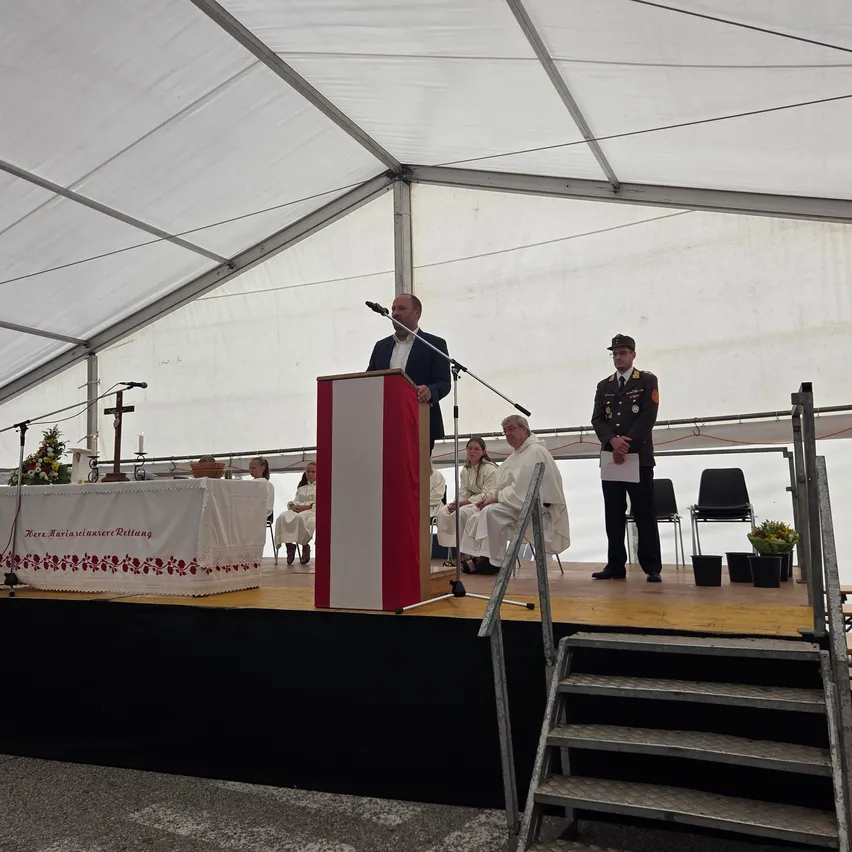 Ein Mann spricht an einem Podium unter einem weißen Zelt, mit einem Militäroffizier und einem sitzenden Priester in der Nähe.