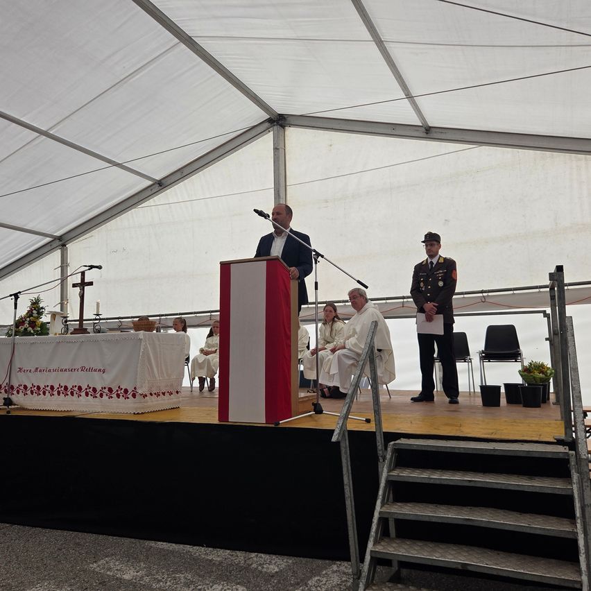 A man speaks at a podium under a white tent, with a military officer and a seated priest nearby.