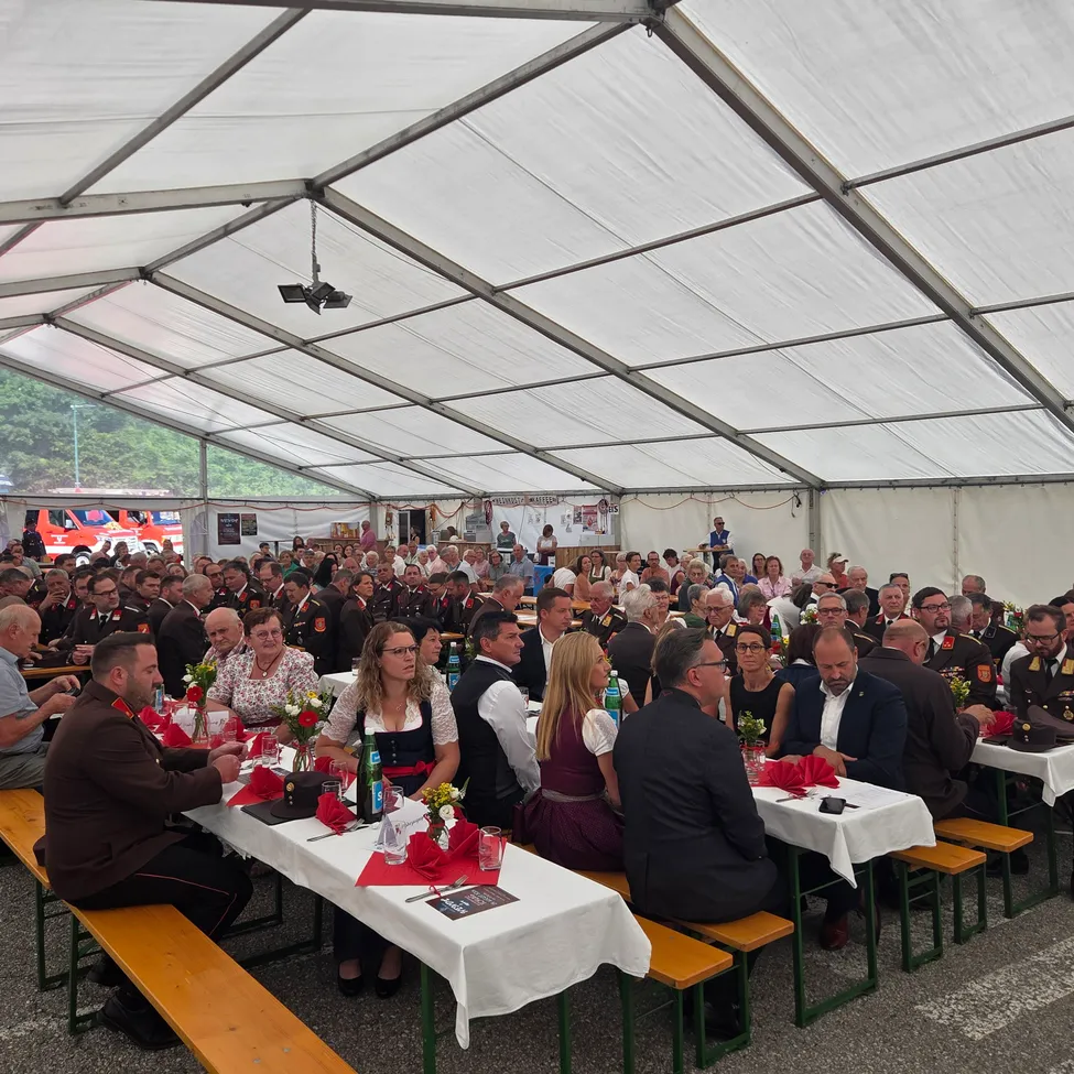 Eine große Gruppe von Menschen sitzt unter einem weißen Zelt an Tischen, möglicherweise bei einer formellen Veranstaltung. Die Tische sind mit weißen Tüchern bedeckt und haben verschiedene Gegenstände darauf, einschließlich Blumen, Flaschen und Servietten. Die Menschen sind in formeller Kleidung gekleidet, und einige tragen Hüte. Im Hintergrund befindet sich ein rotes Fahrzeug und ein grüner Baum.