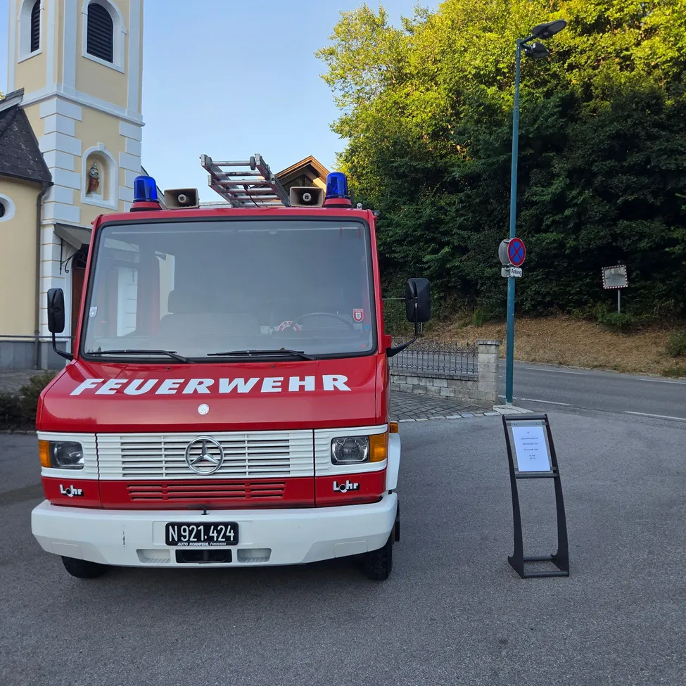 Ein roter Feuerwehrwagen mit dem Wort 'Feuerwehr' auf der Front ist vor einer Kirche geparkt. Eine Straßenlaterne und ein Schild sind in der Nähe.