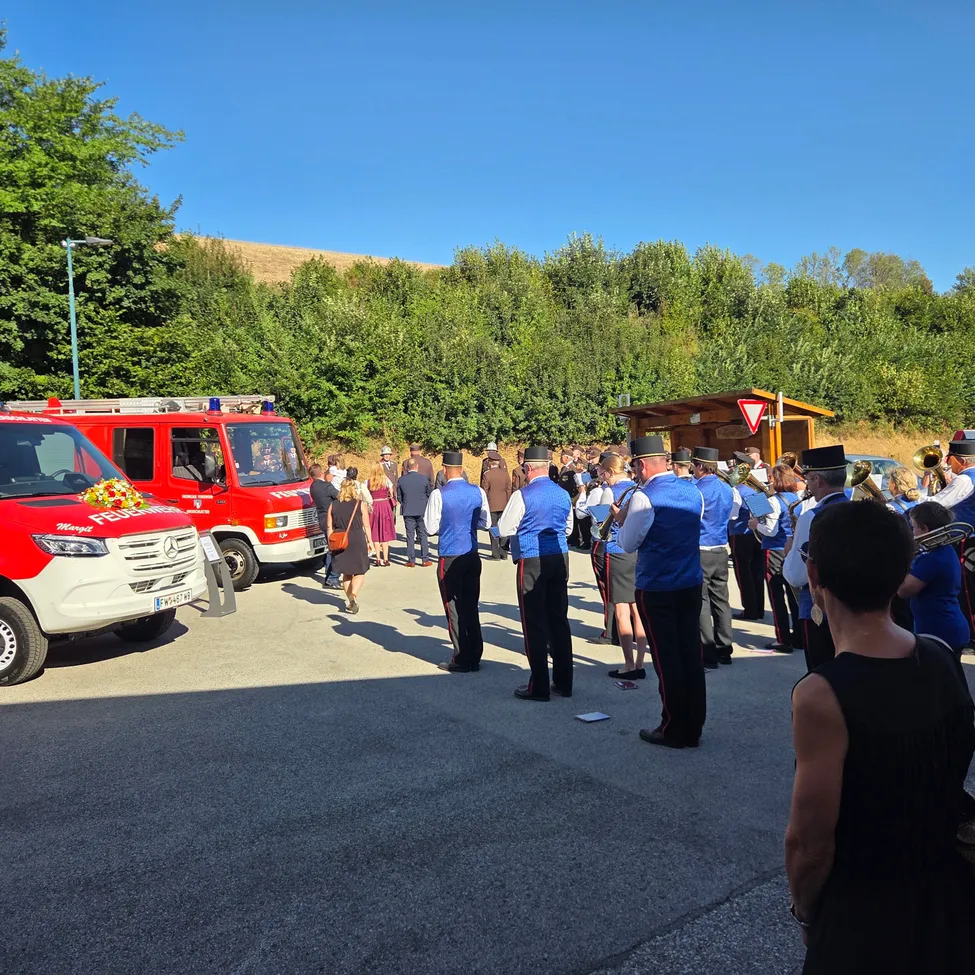 Eine Gruppe von Menschen in blauen Uniformen und Hüten steht in einer Reihe, möglicherweise bei einer Zeremonie. Ein roter Feuerwehrwagen ist auf der linken Seite geparkt. Eine Frau in einem schwarzen Kleid geht in der Mitte. Eine kleine Holzhütte befindet sich auf der rechten Seite.