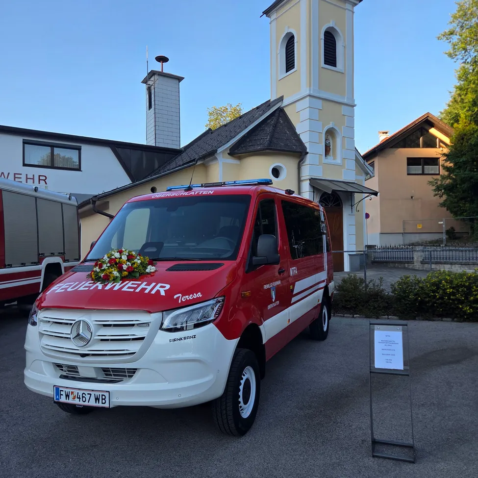 Ein roter und weißer Mercedes-Benz-Van mit dem Wort 'Feuerwehr' auf der Front ist vor einer Kirche geparkt. Der Van hat eine Blumendekoration auf seiner Motorhaube.