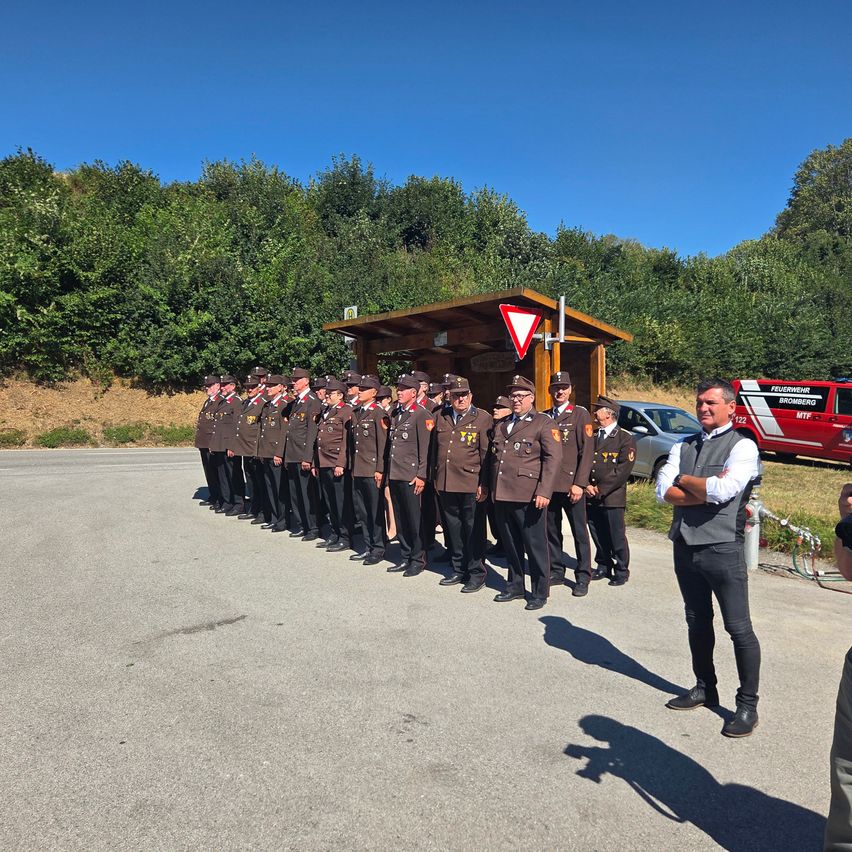 A group of uniformed individuals stands in a row, possibly for a ceremony, with a man standing separately on the right. Behind them, a small shed and a red vehicle are visible.
