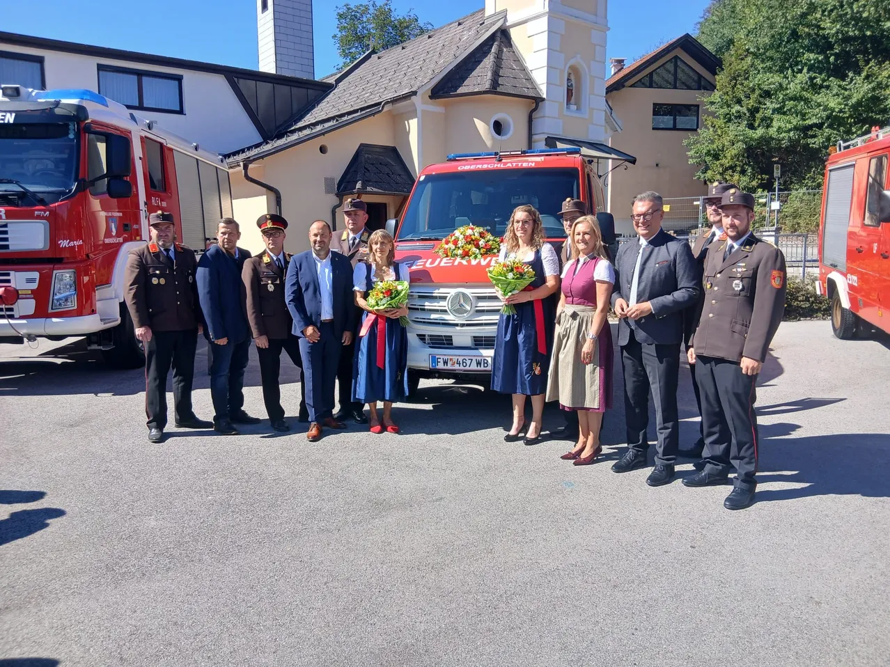 Eine Gruppe von Menschen, einschließlich Beamter und Frauen in traditioneller Kleidung, steht vor einem Feuerwehrauto mit Blumen.