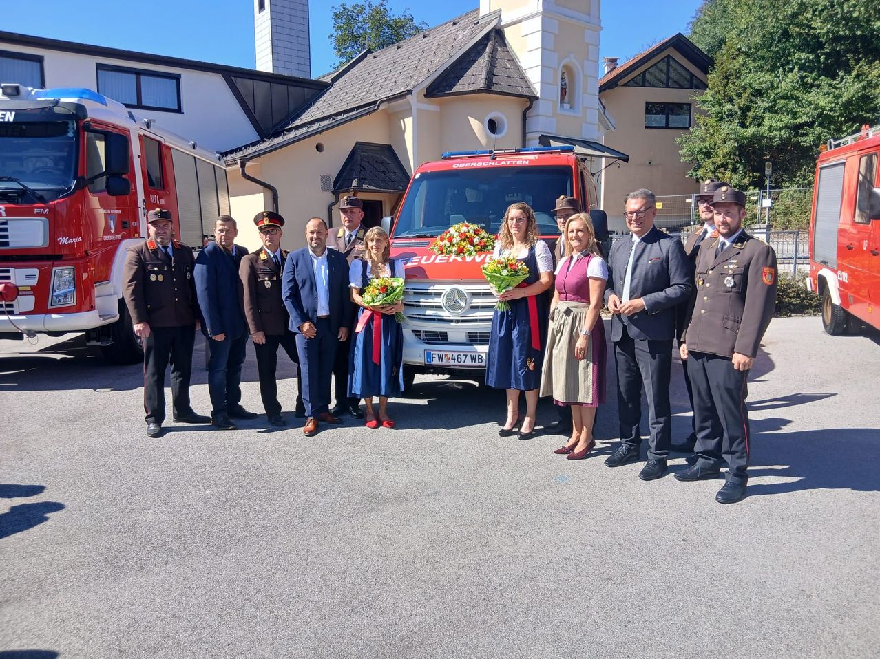 A group of people, including officials and women in traditional attire, stand in front of a fire truck with flowers.
