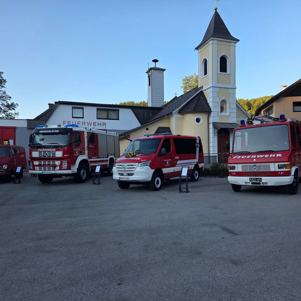 Drei Feuerwehrfahrzeuge sind vor einer Kirche mit einem Turm und einem Kreuz auf der Spitze geparkt. Die Fahrzeuge sind rot und weiß.