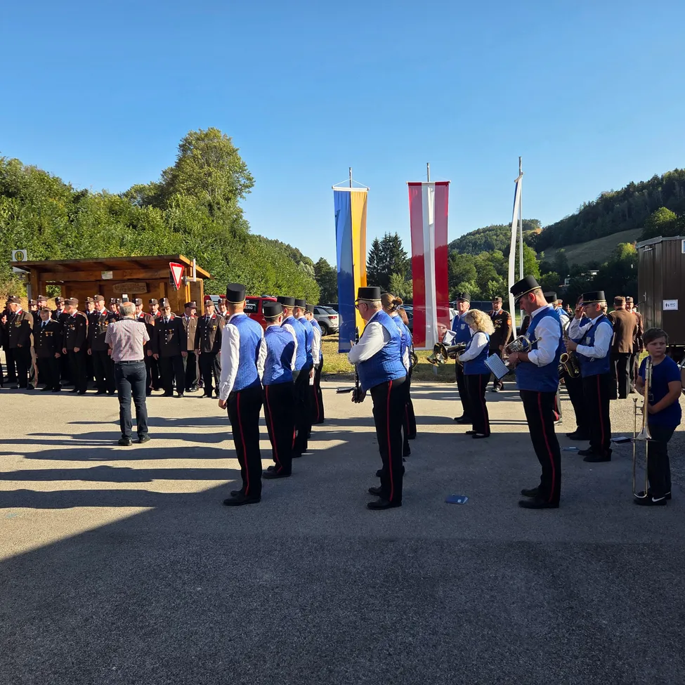 Eine Marschkapelle steht in Formation, Musiker halten Instrumente und Zuschauer beobachten. Der Ort ist im Freien mit Fahnen und Bäumen im Hintergrund.