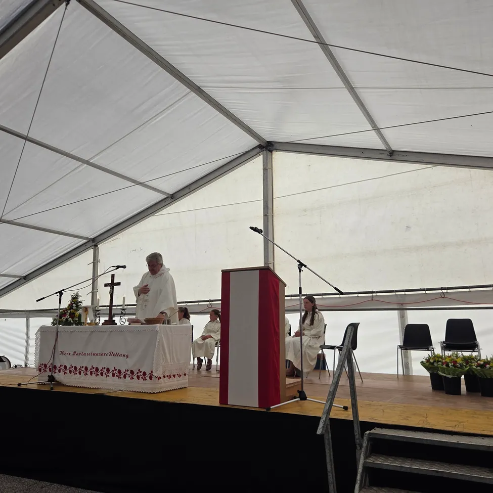 Ein Priester steht an einem Rednerpult in einem Zelt und hält eine Rede. Eine Frau sitzt in einem Stuhl in der Nähe.