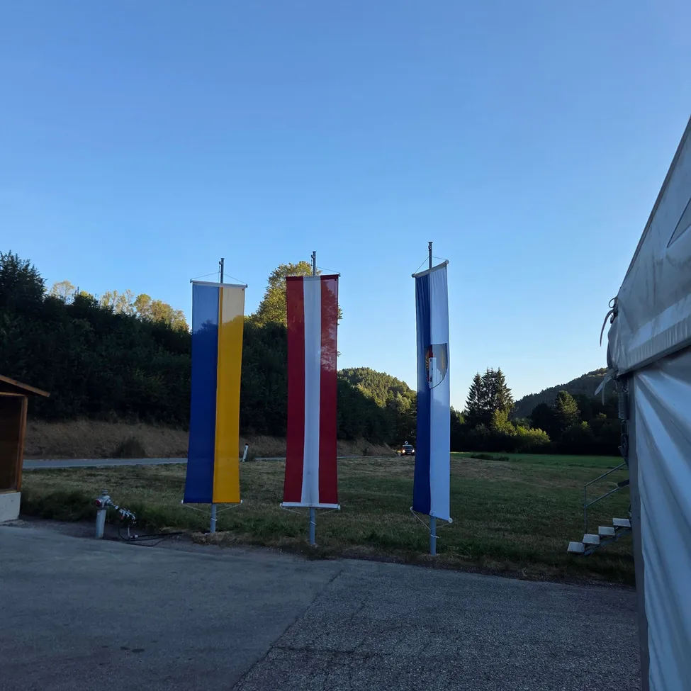 Drei Flaggen, blau und gelb, rot und weiß, und blau und weiß, sind auf Stangen in einem grasigen Feld mit klarem blauem Himmel ausgestellt.