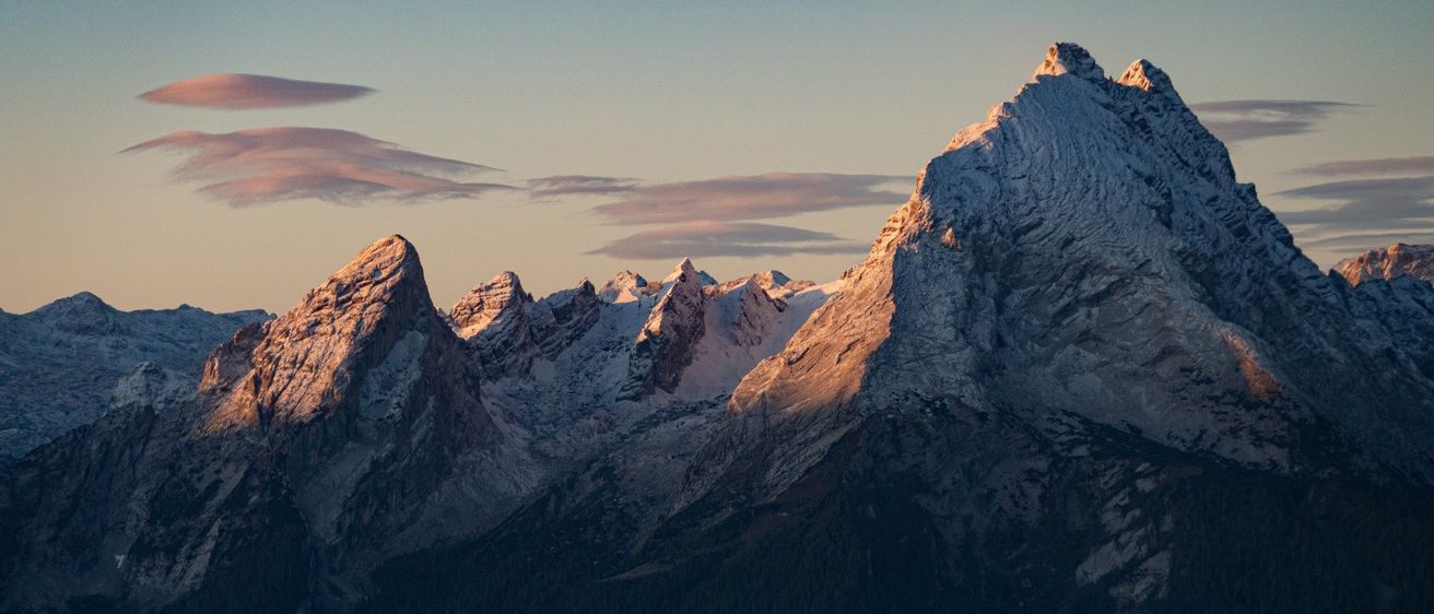 Eine majestätische Bergkette wird vom warmen Licht der untergehenden Sonne beschienen. Schneebedeckte Gipfel und raues Gelände bilden eine atemberaubende Landschaft.