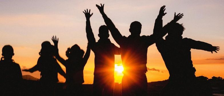 Silhouetten von Menschen, die bei Sonnenuntergang stehen und ihre Hände heben. Die Sonne geht hinter ihnen unter.
