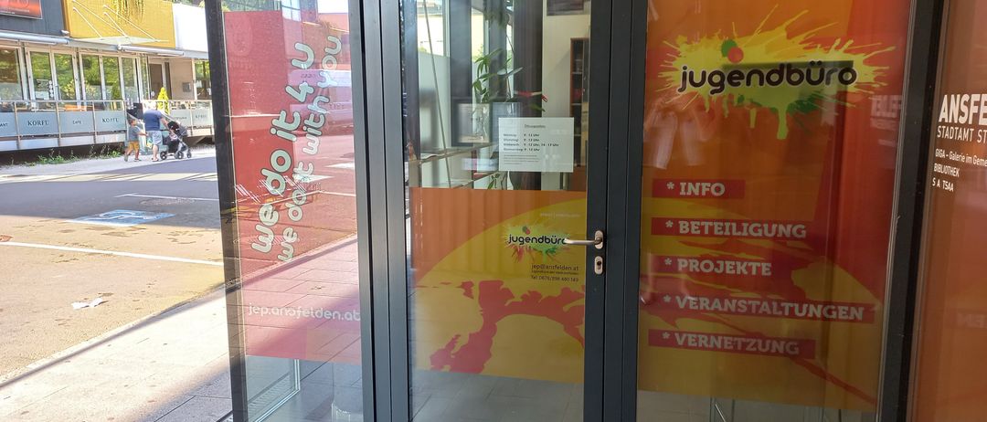 Der Eingang zu einem Gebäude mit Glastüren hat ein leuchtend rotes und oranges Plakat mit weißen Text, der 'jugendbüro' und andere Wörter sagt. Die Türen haben Glasscheiben und einen schwarzen Rahmen.