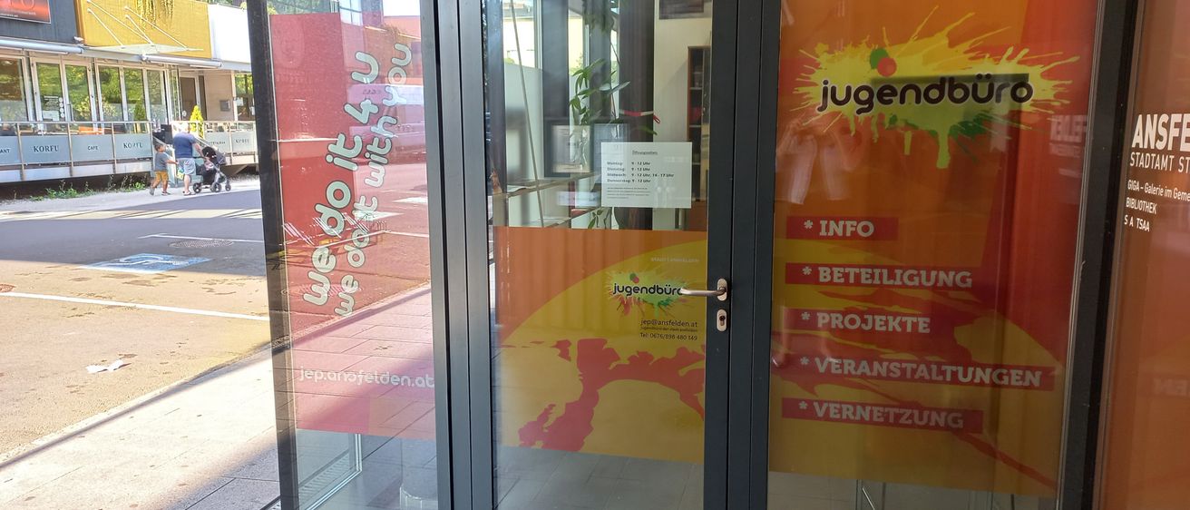 Der Eingang zu einem Gebäude mit Glastüren hat ein leuchtend rotes und oranges Plakat mit weißen Text, der 'jugendbüro' und andere Wörter sagt. Die Türen haben Glasscheiben und einen schwarzen Rahmen.
