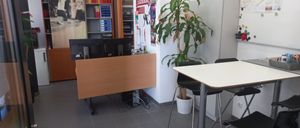 Ein Büro mit einem Schreibtisch, Monitoren, einem Topf mit Pflanze und einer Tafel. Ein Bücherregal und ein Schrank befinden sich im Hintergrund.