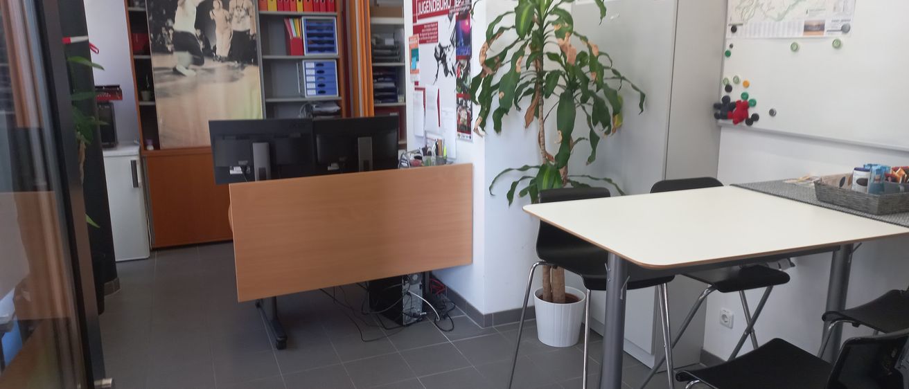 Ein Büro mit einem Schreibtisch, Monitoren, einem Topf mit Pflanze und einer Tafel. Ein Bücherregal und ein Schrank befinden sich im Hintergrund.