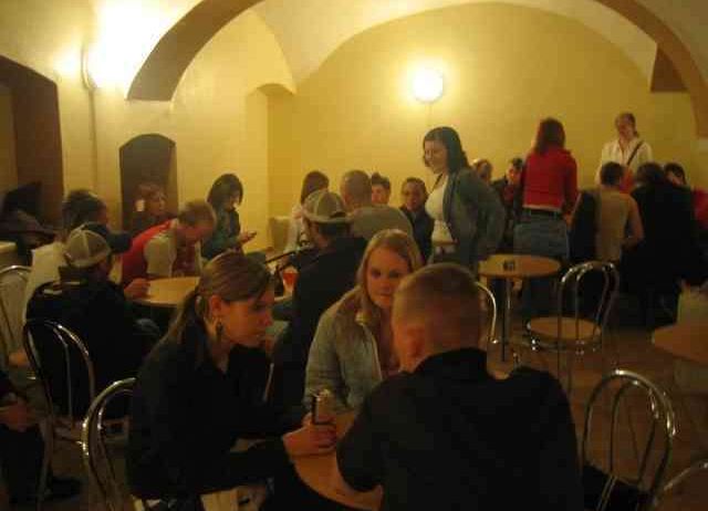 Eine Gruppe von Menschen sitzt an Tischen in einem schwach beleuchteten Raum, einige stehen. Eine Person hält eine Dose. Der Raum hat gewölbte Decken und gelbe Wände.