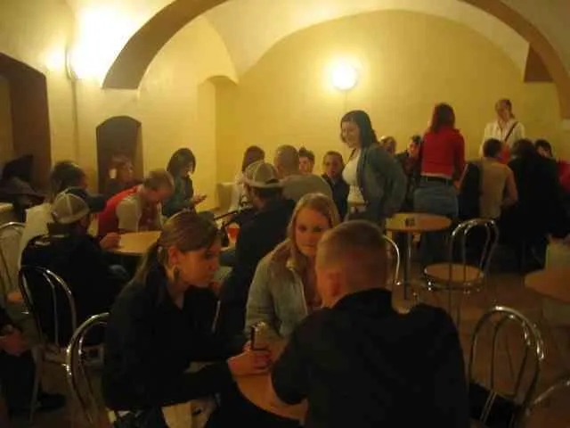 Eine Gruppe von Menschen sitzt an Tischen in einem schwach beleuchteten Raum, einige stehen. Eine Person hält eine Dose. Der Raum hat gewölbte Decken und gelbe Wände.
