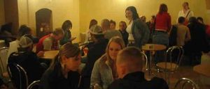 Eine Gruppe von Menschen sitzt an Tischen in einem schwach beleuchteten Raum, einige stehen. Eine Person hält eine Dose. Der Raum hat gewölbte Decken und gelbe Wände.