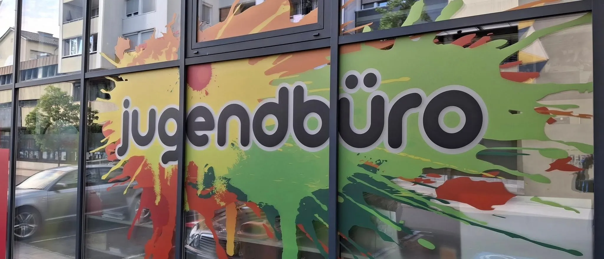 Ein farbenfrohes Schaufenster zeigt 'jugendbuero' in einem lebhaften Farbspritzer-Design, mit Reflexionen eines Gebäudes und Autos im Glas.