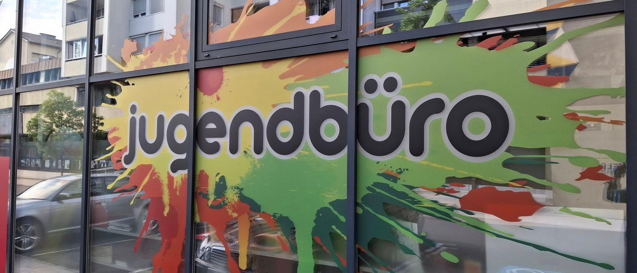 Ein farbenfrohes Schaufenster zeigt 'jugendbuero' in einem lebhaften Farbspritzer-Design, mit Reflexionen eines Gebäudes und Autos im Glas.