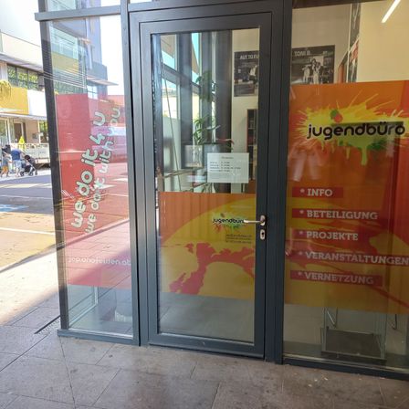 Der Eingang zu einem Jugendzentrum mit einer Glastür, auf der rote und gelbe Schilder angebracht sind. Die Schilder lauten 'Wir machen es gemeinsam' und 'Jugendbüro' und bieten Informationen, Beteiligung, Projekte, Veranstaltungen und Vernetzung an.