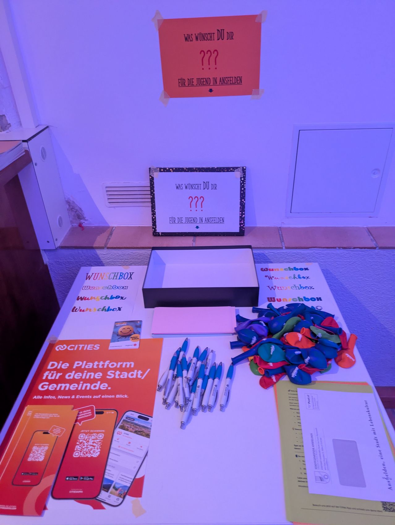 Ein Tisch mit verschiedenen Gegenständen wie Stiften, Papieren und einer Wunschbox. Ein Schild an der Wand fragt: 'Was wünscht du dir?' mit einem Fragezeichen.