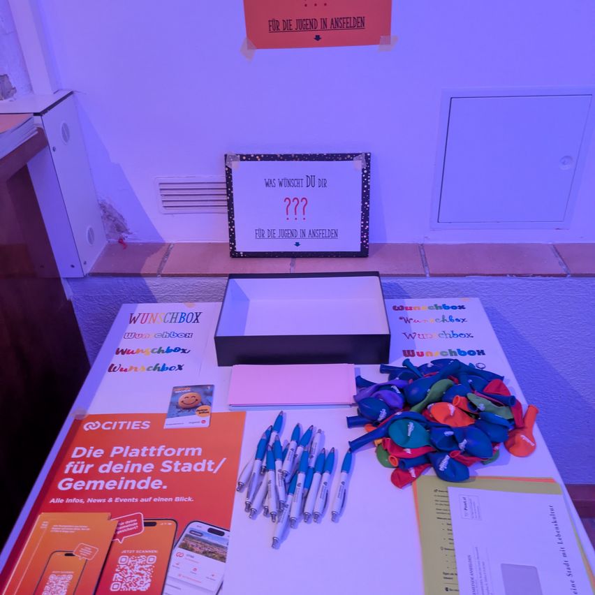 Ein Tisch mit verschiedenen Gegenständen wie Stiften, Papieren und einer Wunschbox. Ein Schild an der Wand fragt: 'Was wünscht du dir?' mit einem Fragezeichen.