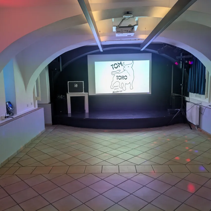 Ein leerer Raum mit einer Bühne und einer Projektionsleinwand, auf der das Wort Toro steht. Die Bühne hat einen weißen Tisch und eine Box.