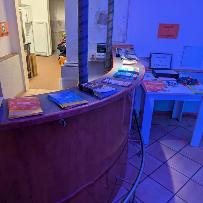 Ein Empfangstresen mit einem lila Schreibtisch hält mehrere Broschüren. Ein weißer Tisch hält zusätzliche Broschüren. Im Hintergrund ist ein Kühlschrank zu sehen. Die Wand hat Schilder mit der Nummer 777.