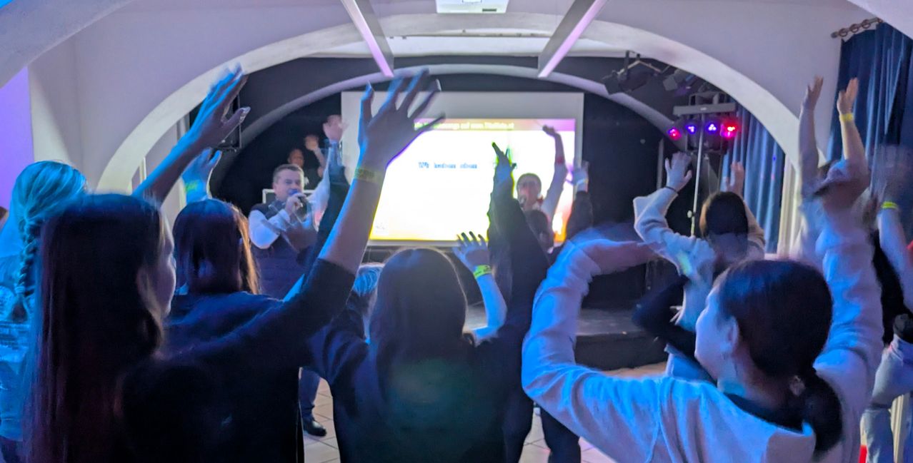 Eine Gruppe von Menschen hebt in einem schwach beleuchteten Raum mit einer Projektionsleinwand und blauen Lichtern die Hände. Ein Mann in einem Anzug steht auf der Bühne und singt in ein Mikrofon.