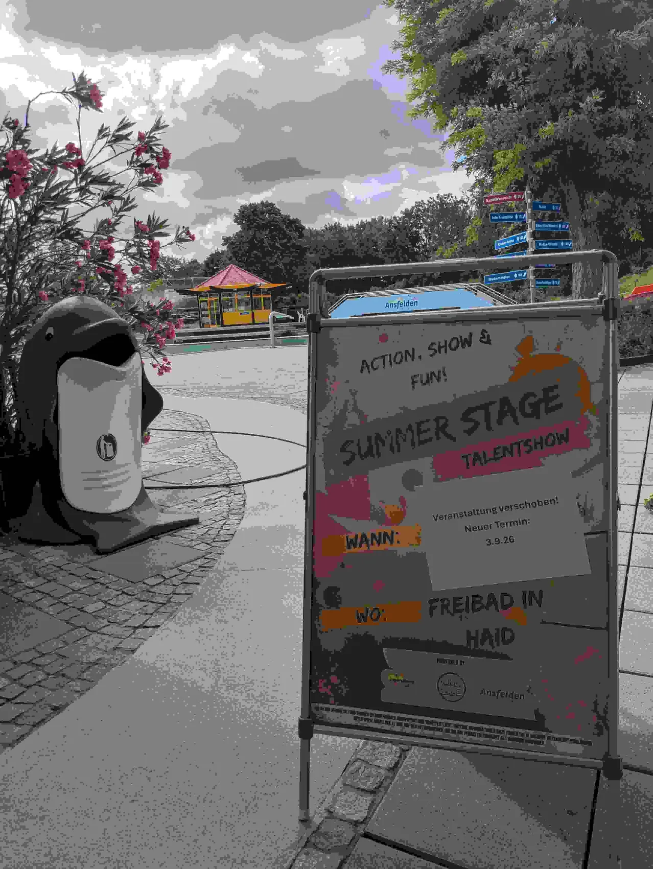 Ein Schild für ein Sommer-Bühnen-Talentshow steht draußen auf einem Ständer. Eine Delfinstatue ist in der Nähe. Das Schild hat ein farbenfrohes Design mit dem Text 'Sommer Stage Talentshow'. Der Hintergrund zeigt einen Weg mit einem kleinen Gebäude und Bäumen.