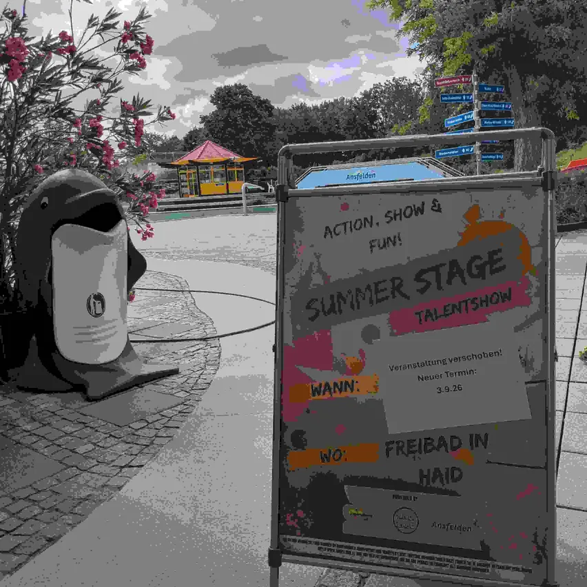 Ein Schild für ein Sommer-Bühnen-Talentshow steht draußen auf einem Ständer. Eine Delfinstatue ist in der Nähe. Das Schild hat ein farbenfrohes Design mit dem Text 'Sommer Stage Talentshow'. Der Hintergrund zeigt einen Weg mit einem kleinen Gebäude und Bäumen.