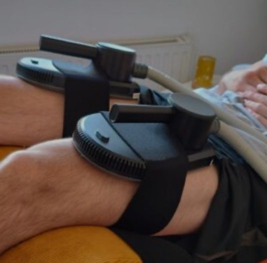 Eine Person liegt mit einem medizinischen Gerät an den Beinen, wahrscheinlich zur Kompressionstherapie. Das Gerät hat einen schwarzen Riemen und eine graue Basis.