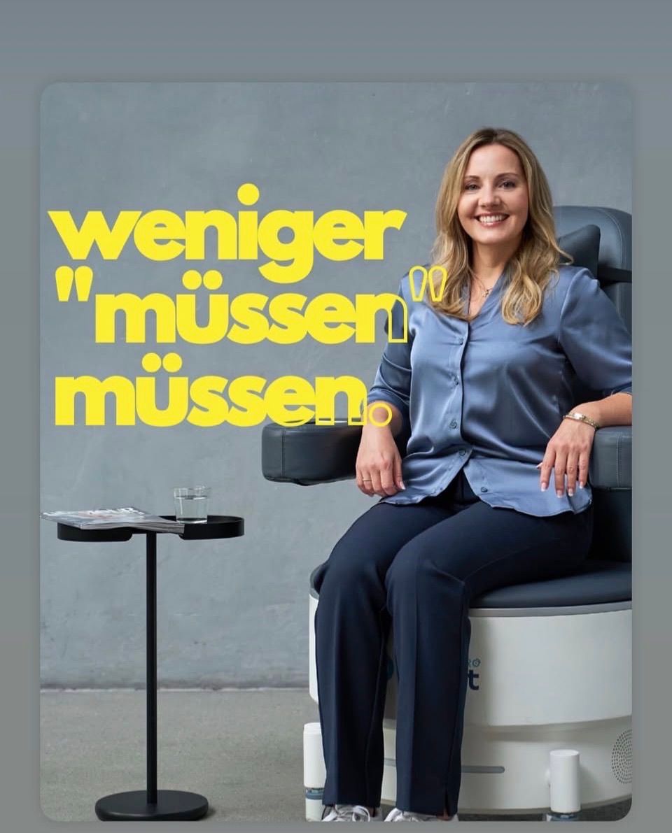 Eine Frau in einem blauen Hemd und Hose sitzt auf einem Stuhl mit einem Glas und einer Zeitschrift auf einem Tisch vor einer grauen Wand mit gelbem Text.