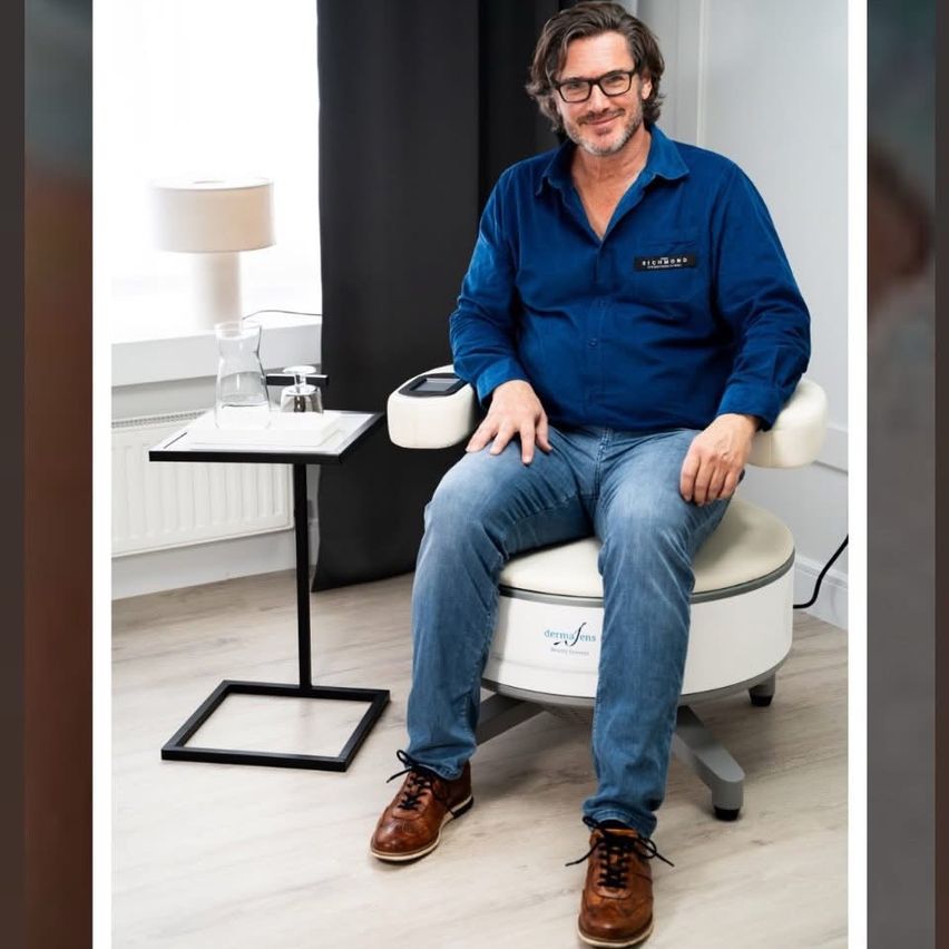 Ein Mann in einem blauen Langarmhemd und Jeans sitzt lächelnd auf einem weißen Stuhl in einem Raum. Hinter ihm steht ein kleiner Tisch mit einer Lampe, einer Glaskaraffe und einem Heizkörper.