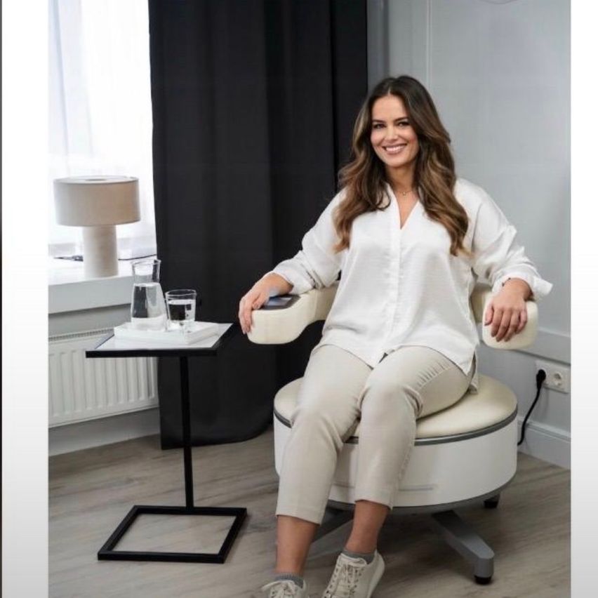 Eine Frau in einem weißen Hemd und Hosen sitzt auf einem weißen Bürostuhl und lächelt. Es gibt einen kleinen Tisch mit einer Lampe und einem Glas Wasser. Hinter ihr ist ein schwarzer Vorhang.