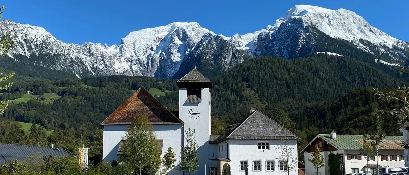 Eine weiße Kirche mit Turm, Uhr und schiefergedecktem Dach steht vor einer schneebedeckten Bergkette.